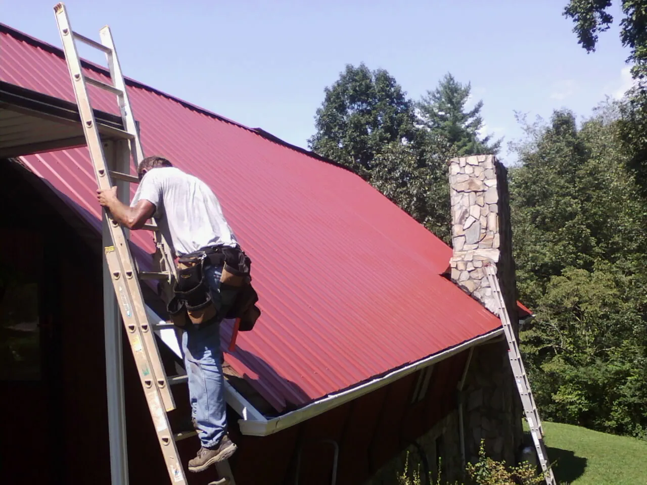 Roofer on a ladder beside a red metal roof and stone chimney — Soffit & Fascia Repair by Roofing Brothers in Hawk Run